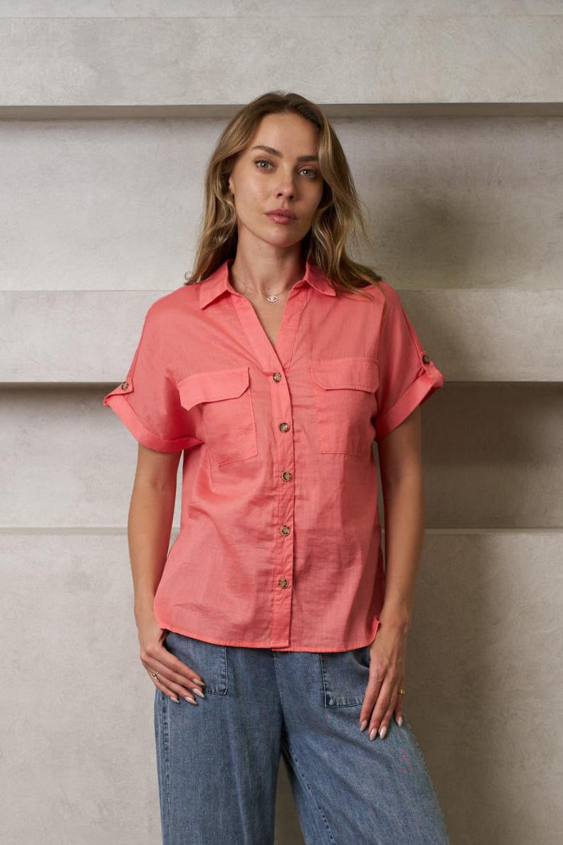 Woman wearing a pink shirt and blue jeans standing against a plain wall.