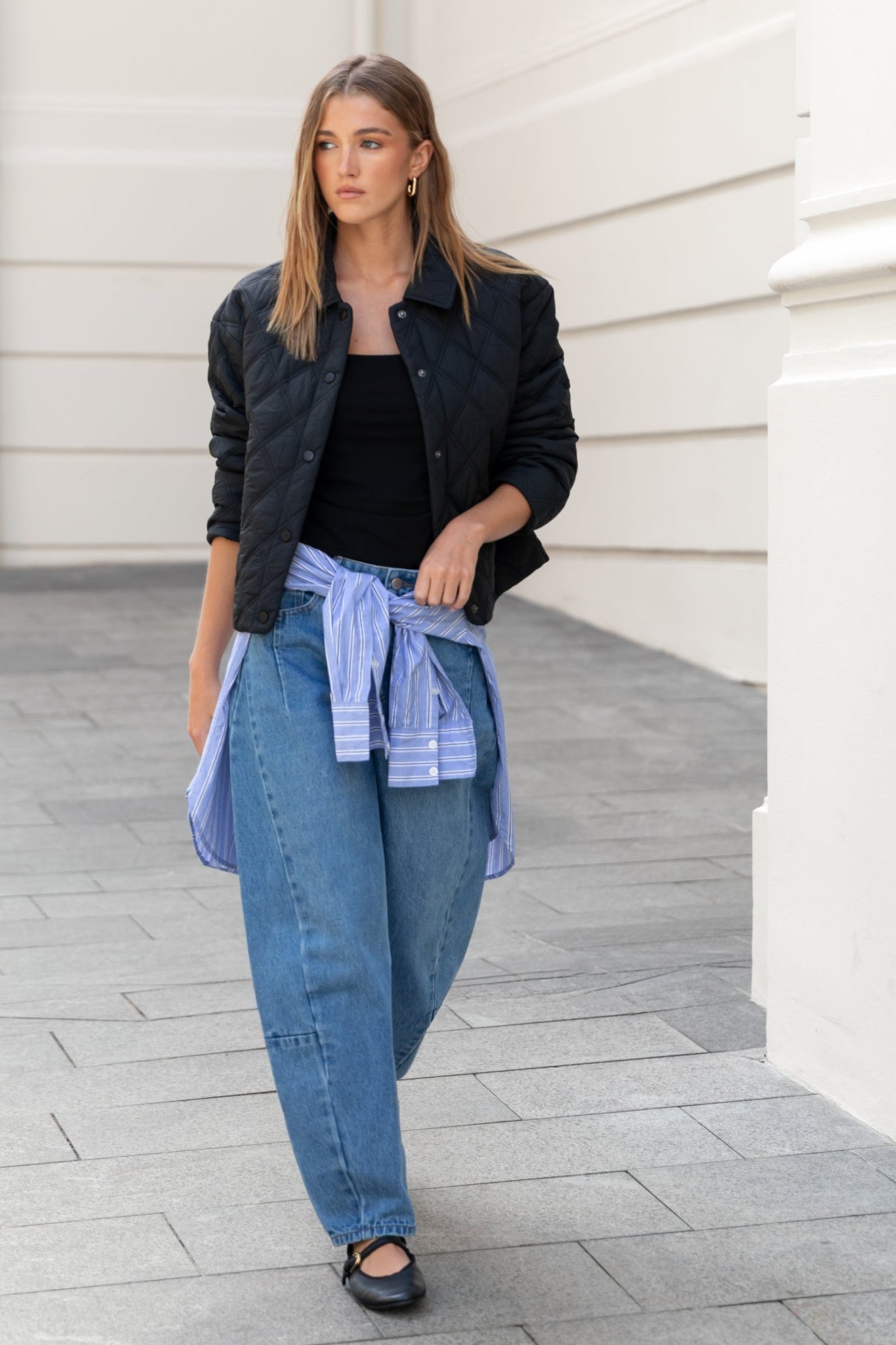Woman walking in an urban setting wearing a black jacket, black top, blue jeans, and sandals.